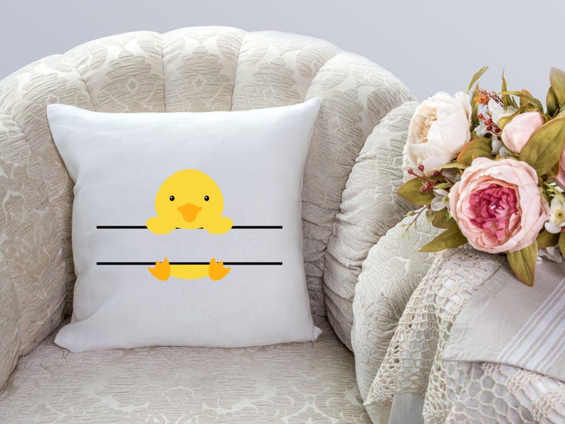May include: White pillow with a yellow duckling peeking over a black line. The duckling has a black beak and orange feet. The pillow is on a cream-colored couch.