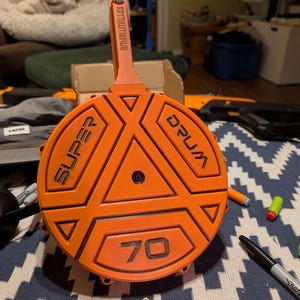 Op de afbeelding: Een oranje "SUPER DRUM 70" magazijn met zwarte letters. Het magazijn heeft een cirkelvormig ontwerp met geometrische vormen en een handvat. De tekst "KPTAUTEKUB" staat op het handvat. Het magazijn is ontworpen voor speelgoedpistolen.