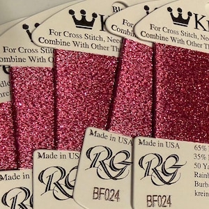 May include: Close-up of several spools of metallic pink thread for cross-stitch and needlepoint. The thread is labeled "Kreinik" and "Made in USA". Each spool contains 50 yards of thread.
