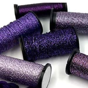 May include: Close-up of several spools of purple and silver metallic thread. The spools are various shades of purple, from deep violet to shimmering silver. The thread is tightly wound on black spools, creating a textured, reflective surface. Ideal for crafting and embroidery projects.