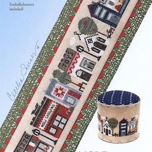 May include: A cross-stitch needlepoint pattern featuring a town scene with buildings, trees, and the text "MORE Any Town tiny town." The design includes a matching pin cushion. The Heart in Hand needleart design is on a beige background.