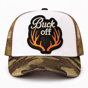 以下が含まれることがあります： 白いフロントパネルと迷彩柄のつばが付いたトラッカーハット。帽子には「Buck off」の文字とオレンジ色の鹿の角が入った黒いパッチが付いています。メッシュの背面は茶色です。
