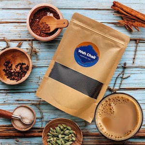 May include: A brown paper bag with a black tea blend inside. The bag has a blue and white logo that says "Wah Chai" Premium Tea. The bag is surrounded by various spices, including cloves, cardamom pods, and cinnamon sticks. A glass of chai tea is also visible.