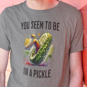 May include: A gray t-shirt with a humorous graphic of a pickle with a person riding it. The text on the shirt reads "You seem to be in a pickle."