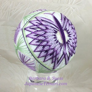 Könnte beinhalten: Eine weiße japanische Temari-Kugel mit einem violetten und grünen Blumenmuster. Die Kugel steht auf einem durchsichtigen Ständer und der Text "Barbara B. Suess JapaneseTemari.com" befindet sich unter der Kugel.