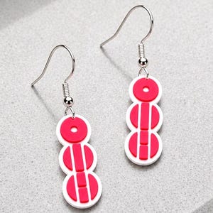 May include: A pair of drop earrings with a white base and red detailing. Each earring has three stacked, circular shapes with a red design. The earrings have silver-coloured hooks.