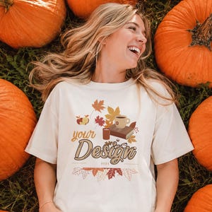 May include: A cream-colored short-sleeve t-shirt featuring a graphic print of autumn leaves, a book, a mug, and an apple. The text "your Design" is printed in a brown script font across the center, surrounded by fall-themed illustrations.