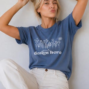 May include: A blue short-sleeved t-shirt featuring a white line-art illustration of various wildflowers across the chest. Below the floral graphic, the text "your design here" is printed in a white, rounded, lowercase sans-serif typeface.