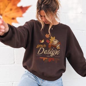 Peut inclure: Un sweat-shirt à col rond marron foncé avec une impression graphique de feuilles d'automne, un livre, une tasse et une pomme. Le texte "your Design HERE" est imprimé au centre du motif. Le sweat-shirt est porté avec un jean bleu clair.