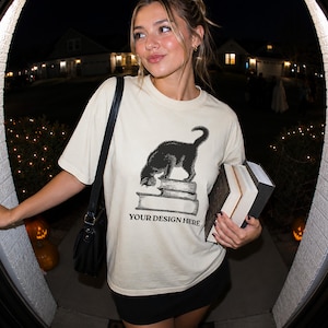 May include: A cream-colored short-sleeve t-shirt featuring a black graphic illustration of a cat standing on a stack of books. The text "YOUR DESIGN HERE" is printed in black capital letters directly below the stack of books on the front.