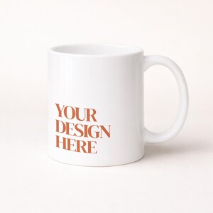 Może przedstawiać: Biały ceramiczny kubek z zakrzywionym uchwytem. Kubek zawiera tekst "YOUR DESIGN HERE" w czerwonobrązowym kolorze. Kubek ma standardowy rozmiar i kształt, odpowiedni do gorących lub zimnych napojów.