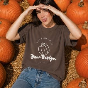 May include: A dark gray short-sleeve t-shirt featuring a white line-art pumpkin graphic. The text "Your Design Here" is printed in a curved arc above the pumpkin, and the words "Your Design" appear in a large script font below it.