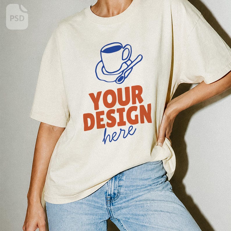 May include: A cream-colored short-sleeve t-shirt featuring a blue graphic of a coffee cup on a saucer with a spoon. Below the graphic, the text reads YOUR DESIGN in bold orange letters, with the word here written in blue cursive script below.