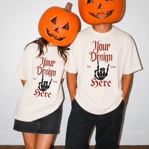 Puede incluir: Dos camisetas de manga corta color hueso con una fuente gótica de color rojo oscuro que dice Your Design Here, junto con el texto Est 2026 y un gráfico de una mano esquelética negra haciendo un gesto de rock en el centro.