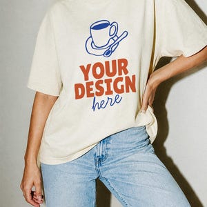 May include: A cream-colored short-sleeve t-shirt featuring a blue graphic of a coffee cup on a saucer with a spoon. Below the graphic, the text reads "YOUR DESIGN" in bold orange letters, followed by the word "here" in blue cursive script.
