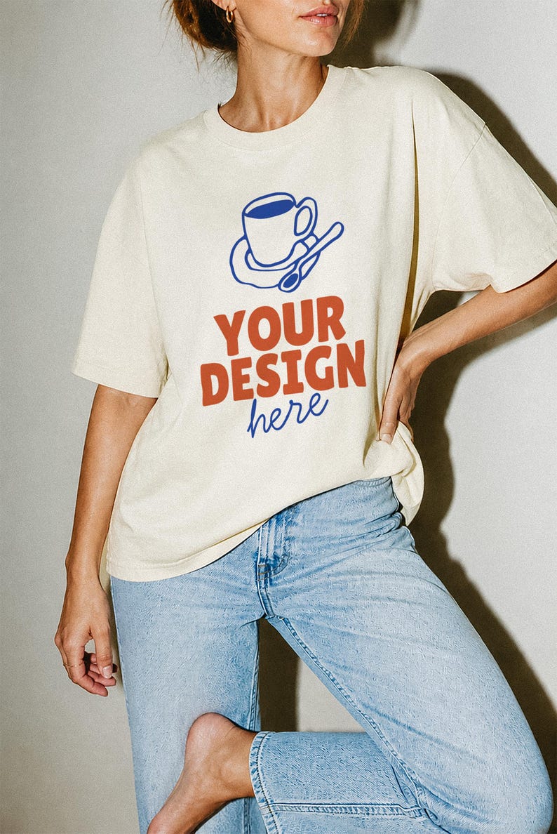 May include: A cream-colored short-sleeve t-shirt featuring a blue graphic of a coffee cup on a saucer with a spoon. Below the graphic, the text reads "YOUR DESIGN" in bold orange letters, followed by the word "here" in blue cursive script.