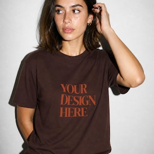 Könnte beinhalten: Ein braunes T-Shirt mit dem Text "YOUR DESIGN HERE" in Orange. Das Shirt ist einfarbig und hat einen Rundhalsausschnitt. Das Model trägt eine blaue Jeans.