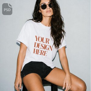 Może przedstawiać: Biały t-shirt z krótkim rękawem z napisem YOUR DESIGN HERE wydrukowanym rdzawą czcionką szeryfową na klatce piersiowej. Koszulka ma okrągły dekolt i podwinięte rękawy, zestawiona z czarnymi szortami. Prezentowana jako szablon typu mockup.