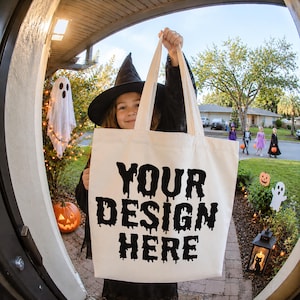 May include: A cream-colored canvas tote bag featuring the black text YOUR DESIGN HERE in a dripping, spooky font style. The bag has two long fabric handles and is held up against a background showing a person in a witch costume.