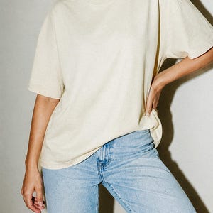 May include: A short-sleeved, crew-neck t-shirt in a solid cream color paired with light-wash blue denim jeans. The t-shirt features a relaxed, loose-fitting silhouette with a standard hemline, while the jeans have a classic five-pocket design.