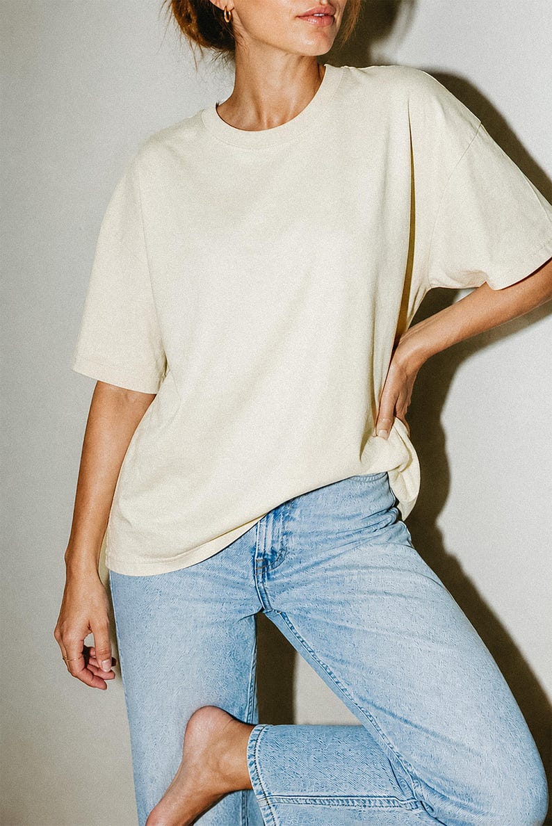 May include: A short-sleeved, crew-neck t-shirt in a solid cream color paired with light-wash blue denim jeans. The t-shirt features a relaxed, loose-fitting silhouette with a standard hemline, while the jeans have a classic five-pocket design.