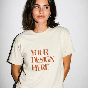May include: A cream-colored short-sleeve t-shirt featuring the text "YOUR DESIGN HERE" printed in a bold, rust-colored serif font across the chest. The garment has a classic crew neckline and a standard fit, providing a neutral base for customization.