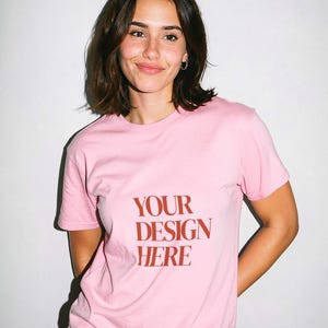 May include: A light pink t-shirt with the text "YOUR DESIGN HERE" in a bold, red font. The shirt is short-sleeved and appears to be made of a soft material. The model is wearing blue jeans.