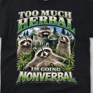 May include: Black t-shirt with a graphic featuring raccoons and the text "TOO MUCH HERBAL I'M GOING NONVERBAL". The design includes raccoons, a metal bin, and green foliage.