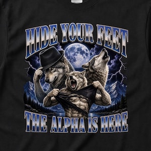 Puede incluir: Una camiseta negra de manga corta con un estampado gráfico de tres lobos bajo una luna llena con rayos y montañas. El texto dice HIDE YOUR FEET y THE ALPHA IS HERE. Un lobo lleva un sombrero fedora y otro se rasga la camisa.