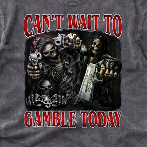 May include: Dark grey t-shirt featuring a graphic of skeletal figures holding guns, with the text "CAN'T WAIT TO GAMBLE TODAY" in red. The design is centered on the front of the shirt, with a vintage wash effect.