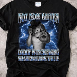 May include: Black t-shirt with a graphic featuring a cartoon wolf wearing a hat, with text that reads "NOT NOW KITTEN" and "DADDY IS INCREASING SHAREHOLDER VALUE" in a blue font. The background has blue lightning bolts.