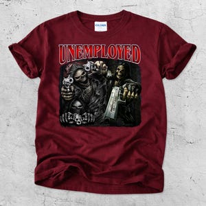 May include: Maroon t-shirt with the word "UNEMPLOYED" in red and white lettering. The graphic depicts a skeletal figure holding a gun, with other skulls and guns. The shirt is made of a soft material.