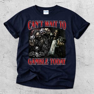 May include: Navy blue t-shirt with a graphic featuring skeletal figures holding guns. The text "CAN'T WAIT TO GAMBLE TODAY" is in red, outlined in white. The shirt is made of a soft material.