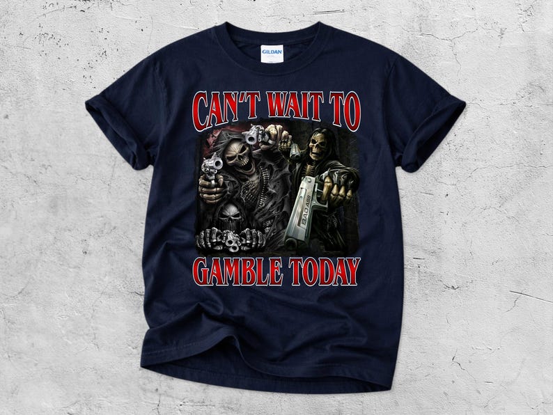 May include: Navy blue t-shirt with a graphic featuring skeletal figures holding guns. The text "CAN'T WAIT TO GAMBLE TODAY" is in red, outlined in white. The shirt is made of a soft material.