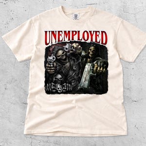 May include: Cream-colored t-shirt featuring a graphic of two skeletal figures holding guns, with the word "UNEMPLOYED" in red above them. The design includes skulls and the text "BAD ASS" on one of the guns.