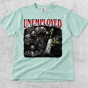 May include: A light blue t-shirt featuring the word "UNEMPLOYED" in red above a graphic of the Grim Reaper holding guns. The graphic is detailed with skulls and dark colors.