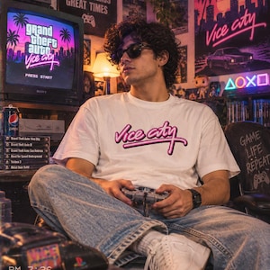 Op de afbeelding: Een wit t-shirt met de roze neon-stijl tekst Vice City op de borst. Het shirt wordt gedragen met een lichtblauwe spijkerbroek, witte sneakers en een zwart digitaal horloge. Het ontwerp weerspiegelt een retro esthetiek met een gaming thema.