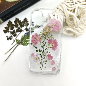 May include: Clear phone case with embedded pink flowers and green stems. The case has a glittery, sparkly appearance. Dried flowers and leaves are visible near the case.