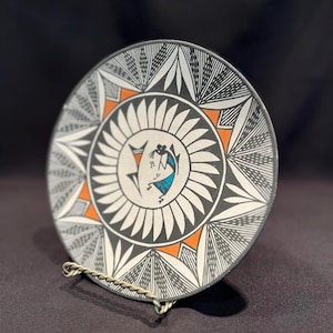 May include: A black and white ceramic plate with a geometric design and a turquoise and orange bird in the center. The plate is on a gold stand.