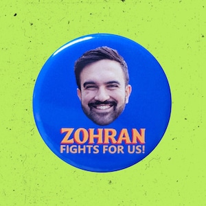 May include: A circular blue pinback button featuring a photographic portrait of a smiling person with dark hair and a beard. Below the portrait, the text ZOHRAN FIGHTS FOR US! is printed in bold, orange, capitalized letters with a dark outline.