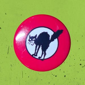 May include: A circular pinback button with a bright red background. In the center, a white circle contains a black silhouette of an arched-back cat with its fur standing up, depicted in a classic hissing pose with a raised, bushy tail.