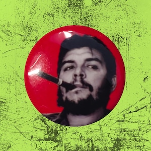 May include: A circular pinback button featuring a black and white photographic portrait of Che Guevara smoking a cigar against a solid red background. The button has a glossy finish and displays the iconic image of the historical figure.