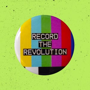 May include: A circular pin-back button featuring a vertical color bar test pattern in yellow, light blue, green, magenta, and red. The text "RECORD THE REVOLUTION" is printed in a bold, black, pixelated font across the center of the design.