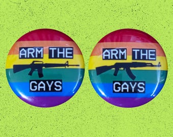 May include: Two circular pin-back buttons featuring a horizontal rainbow flag background. Each button displays the black text ARM THE GAYS in a blocky font, with a black silhouette of a rifle positioned between the two lines of text.