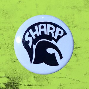 May include: A circular pin-back button featuring a white background with a black graphic of a Spartan helmet. The word SHARP is printed in bold black capital letters across the top of the helmet design, creating a clear and high-contrast visual.
