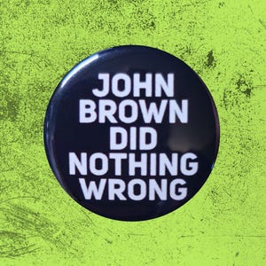 May include: A circular pin-back button with a glossy black surface and bold white sans-serif text that reads JOHN BROWN DID NOTHING WRONG in four stacked lines. The button has a smooth, reflective finish and a clean, simple design.