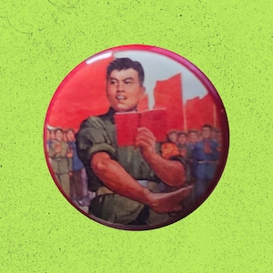 May include: A circular pinback button featuring a graphic illustration of a person in a green uniform holding a small red book. The background depicts a crowd of people and red structures under a bright red sky, rendered in a propaganda art style.