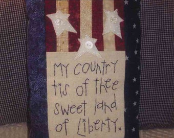 May include: A handmade pillow with a patriotic theme. The pillow is made of blue, red, and white fabric and features a stitched design of stars and stripes. The pillow has a white fabric panel with the words "My country tis of thee sweet land of liberty" stitched in black thread.