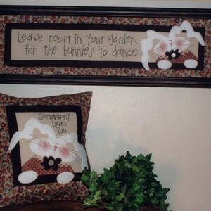 May include: Two framed wall decorations with a bunny theme. The top frame has a floral patterned background with the text "Leave room in your garden for the bunnies to dance". The bottom frame is a pillow with a floral patterned background and the text "Somebody loves you" with a white bunny in the center.