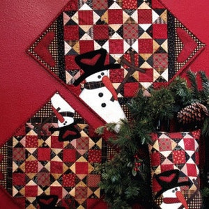 May include: Three quilted wall hangings featuring a snowman design. Each hanging is made of red, black, and white fabric with a patchwork pattern. The hangings are arranged in a triangular shape on a red wall.